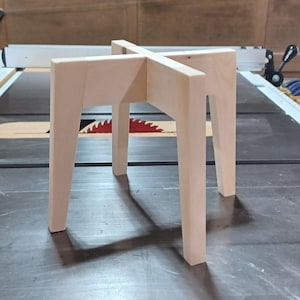 May include: A small, unfinished wooden plant stand with a cross-shaped top and four straight legs. The stand is made of light-colored wood and has a simple, geometric design. The stand is approximately 12 inches tall.