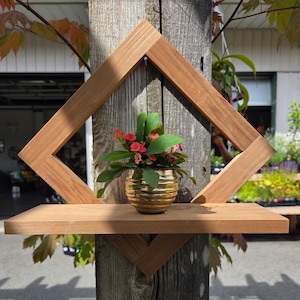 Puede incluir: Estante de madera con marco en forma de diamante, montado en un poste de madera desgastada. Una pequeña planta en maceta con flores rojas y hojas verdes se encuentra en el estante. La madera tiene un acabado marrón claro natural.