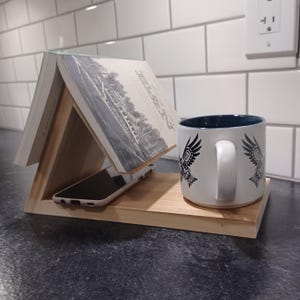 May include: A wooden phone and book stand with a white mug with a black eagle design. The mug is sitting on the stand, and a book is propped up against the stand.