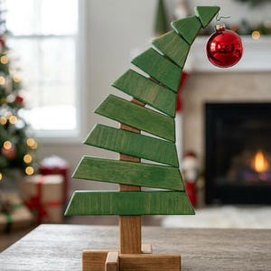 May include: A wooden Christmas tree decoration featuring a natural brown base and a green, slanted, tiered structure. The tree is topped with a single, shiny red spherical ornament hanging from the pointed tip of the wooden frame.