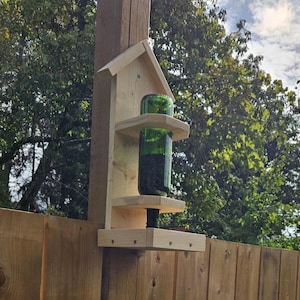 Könnte beinhalten: Ein hölzerner Vogelhäuschen mit einer grünen Glasflasche an der Seite. Der Futterspender ist an einem Holzpfosten befestigt und hat ein kleines Dach.