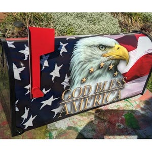 May include: A black mailbox with a red flag. The mailbox is decorated with an American flag design, a bald eagle, and the words "God Bless America".