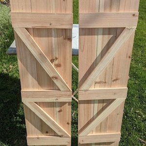 May include: Two unfinished wooden shutters with a diagonal X design. The shutters are made of light brown wood and have a rustic look.