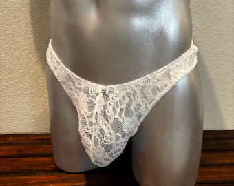May include: A white lace thong featuring a detailed floral pattern throughout the fabric. The garment is constructed from a sheer, delicate material with a scalloped edge along the waistband and leg openings, providing a lightweight and textured appearance.