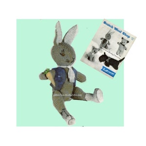 May include: A gray knitted rabbit toy holding a carrot. The rabbit is wearing a blue vest and a white scarf. The toy is sitting on a light green background. The image includes the text "Scotch Wool Shop" and "pattern from WonkyZebra.com".