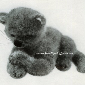 May include: A gray knitted cat toy with a fluffy texture. The cat is sitting with its paws tucked under its chin, looking down. The toy is made with a pattern from WonkyZebra.com.