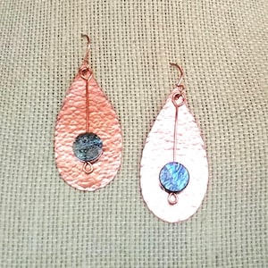 May include: A pair of teardrop-shaped copper earrings featuring a hammered texture. Each earring has a vertical copper wire centered with a small, circular, iridescent blue and purple metallic disc. The earrings hang from simple copper ear wires.