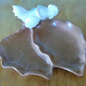 May include: A brown ceramic serving dish shaped like two leaves with a white ceramic rooster figurine on top. The dish has a glossy finish and is divided into two sections.