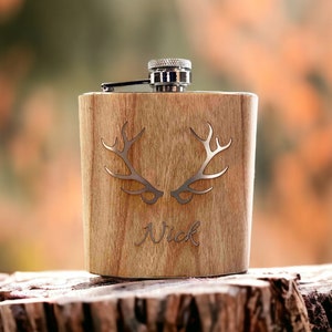 Puede incluir: Una petaca de madera con tapón plateado y cierre de bisagra. Presenta un diseño de cornamenta plateada y el nombre "Nick" grabado en la parte delantera. La petaca está sobre un tocón de árbol.