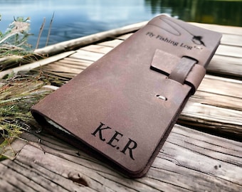 May include: A brown leather fly fishing log with the initials "K.E.R." engraved on the cover. The log features a strap closure and the words "Fly Fishing Log" with a fish graphic. The log is resting on a wooden dock.
