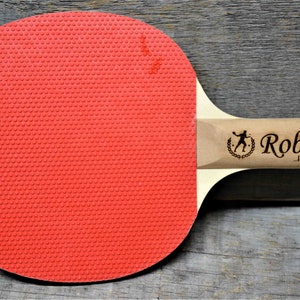 May include: A red and wooden ping pong paddle with the name "Robert" and the text "I own this table" engraved on the handle.