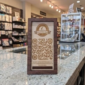 Peut inclure: Un panneau en bois encadré avec un code QR et le texte "MEMORIES MADE". Le panneau est posé sur un comptoir dans un magasin. Des étagères de produits sont visibles en arrière-plan.