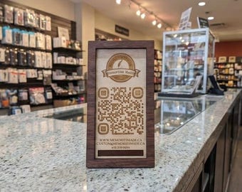 May include: A wooden framed sign with a QR code and the text "MEMORIES MADE". The sign is on a countertop in a store. The store has shelves of products in the background.
