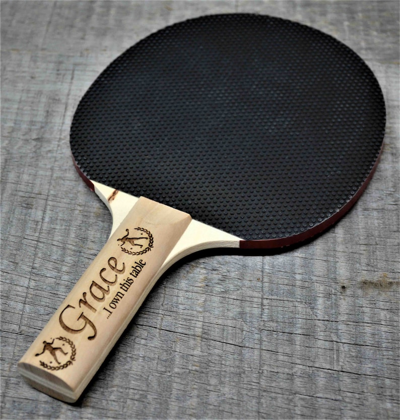 May include: A black and red ping pong paddle with a wooden handle. The handle is engraved with the name "Grace" and the text "I own this table".