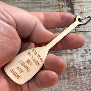 Puede incluir: Un llavero de madera de color claro con forma de remo. El llavero tiene un anillo de metal y el texto "CUSTOM WOODEN KEY CHAINS ANY SHAPE" grabado en la paleta. El llavero se sostiene en una mano.