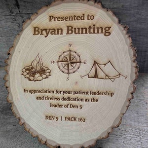 Peut inclure: Une plaque en bois avec un motif de grain de bois, représentant une boussole, un feu de camp et une tente. La plaque indique "Présenté à Bryan Bunting" et "En reconnaissance de votre leadership patient et de votre dévouement indéfectible en tant que chef de la meute 5." La plaque indique également "DEN 5 | PACK 162".