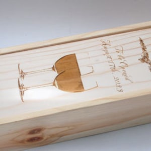 May include: A wooden box with a sliding lid, engraved with the text "To be Opened August 7th, 2023" and "Jill & Neil". The box is lined with brown paper shreds and features a design of two wine glasses.