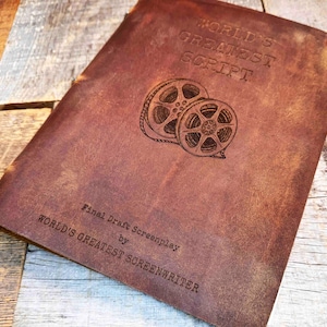 May include: A brown leather-bound notebook with the words "WORLD'S GREATEST SCRIPT" embossed on the cover. Below the text are two film reel illustrations. The bottom of the cover reads "Final Draft Screenplay by WORLD'S GREATEST SCREENWRITER."