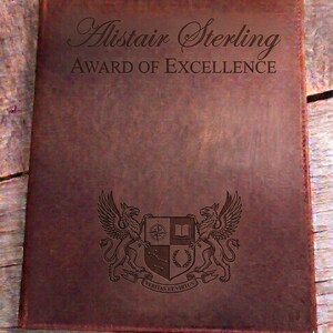 May include: A brown leather award certificate featuring the name "Alistair Sterling" and "AWARD OF EXCELLENCE". The certificate has an engraved crest with two griffins, a compass, a book, and the motto "Veritas et Virtus".