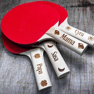 May include: Four red and white ping pong paddles with wooden handles. The handles are personalized with the names "Cyra", "Mama", "Sasa", and "Pappa".