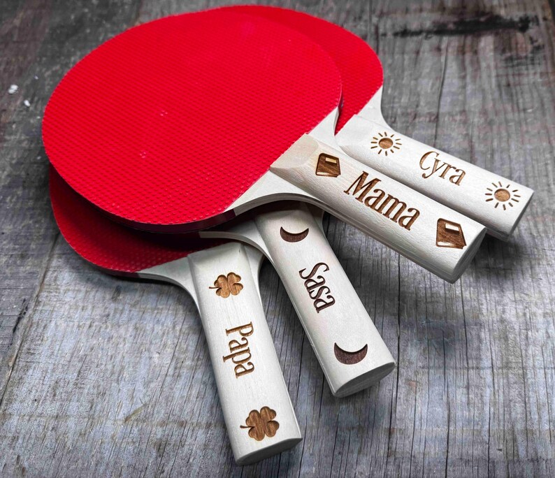 May include: Four red and white ping pong paddles with wooden handles. The handles are personalized with the names "Cyra", "Mama", "Sasa", and "Pappa".