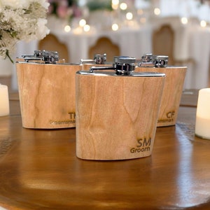 Puede incluir: Cuatro favores de boda de frasco de madera con nombres grabados y las palabras "Groom" y "Groomsman". Los frascos están sobre una mesa de madera con una vela blanca.