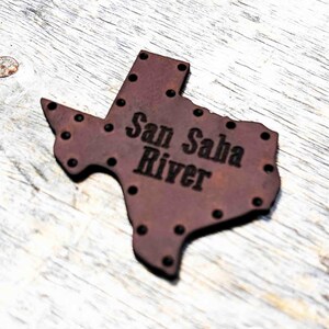 Pode incluir: Um recorte de couro marrom em forma do estado do Texas com as palavras "San Saba River" gravadas nele. O recorte tem pequenos pontos pretos ao redor das bordas.