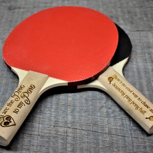May include: Two wooden ping pong paddles with engraved text. The red paddle has the text "You are the DING to my DONG" and the black paddle has the text "There's no cool way to chase after a bouncing ping pong ball."