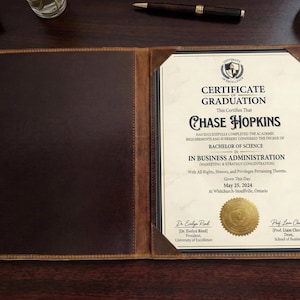 以下が含まれることがあります： 卒業証明書が入った茶色の革製証明書ホルダー。証明書は、2024年5月25日付のChase Hopkins、経営学士号用です。ホルダーは、濃い木製の表面に開いています。