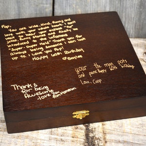Pode incluir: Uma caixa de madeira castanho escuro com uma trava dourada. A parte superior da caixa &eacute; gravada com uma mensagem escrita &agrave; m&atilde;o que diz: "Pop, You are wise, kind, funny and love with your whole heart. I can't wait to hear your endless "Top" jokes, funny stories and finally be introduced to the infamous "Super-Duper Hold" Thank you for always being someone I can look up to. I love you Pop! Happy 60th Birthday Baylee. Thank for being Awesome you Kameron -love from your the man pots and pans. Happy 60th birthday Love, Coop"