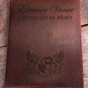 May include: A brown leather certificate holder. The cover features the name "Eleanor Vance" and "CERTIFICATE OF MERIT" in script. A detailed crest with griffins and Latin text is embossed at the bottom. The holder is rectangular.