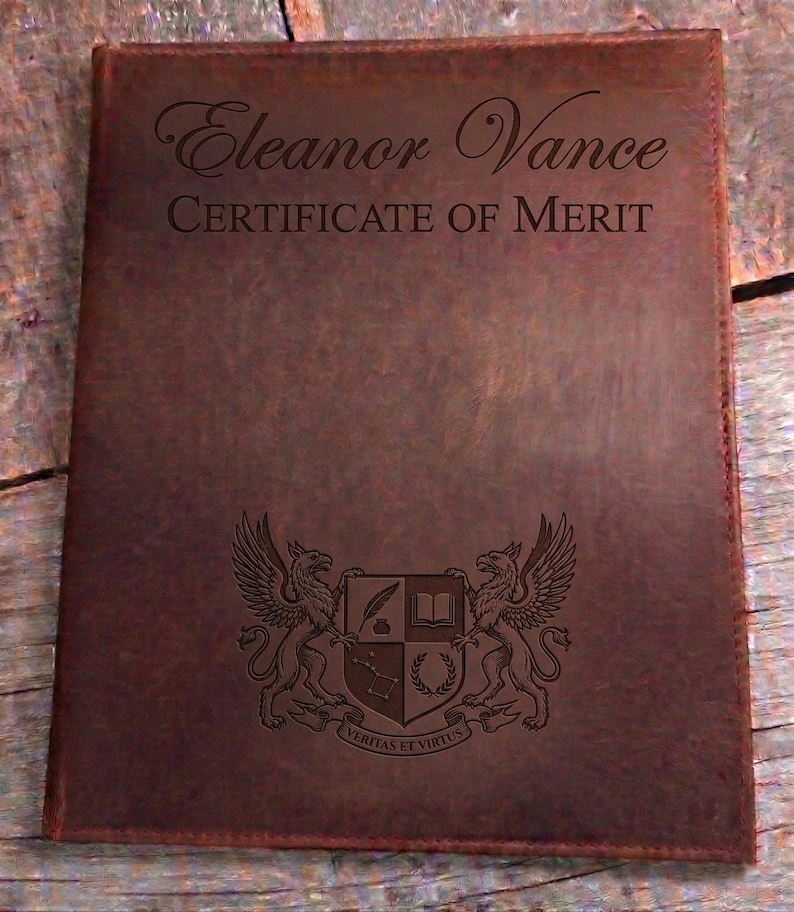 May include: A brown leather certificate holder. The cover features the name "Eleanor Vance" and "CERTIFICATE OF MERIT" in script. A detailed crest with griffins and Latin text is embossed at the bottom. The holder is rectangular.