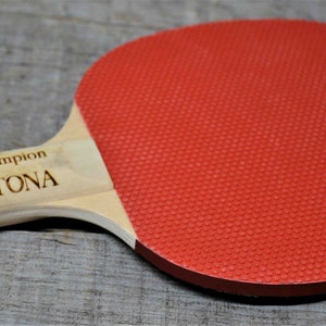 May include: A wooden ping pong paddle with a red rubber surface. The paddle is engraved with the text "Ping Pong Champion Randy Daytona".