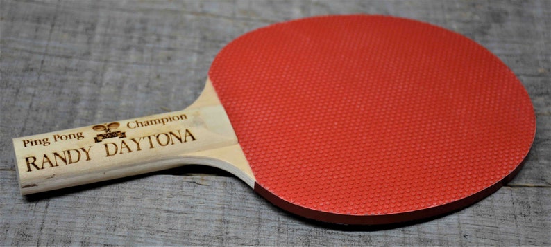 May include: A wooden ping pong paddle with a red rubber surface. The paddle is engraved with the text "Ping Pong Champion Randy Daytona".