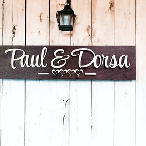 May include: A wooden sign with the words "Paul & Dorsa" in white lettering. The sign is decorated with five small white hearts.