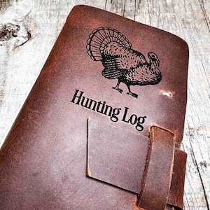 May include: A brown leather hunting log featuring a turkey illustration and the words "Hunting Log" printed in black. The journal has a leather strap closure.