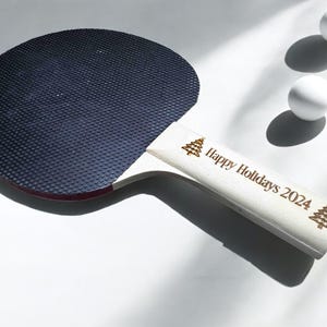 May include: A white ping pong paddle with a black rubber surface and a handle engraved with "Happy Holidays 2024" and two small Christmas tree designs.