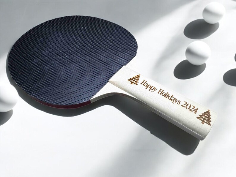 May include: A white ping pong paddle with a black rubber surface and a handle engraved with "Happy Holidays 2024" and two small Christmas tree designs.