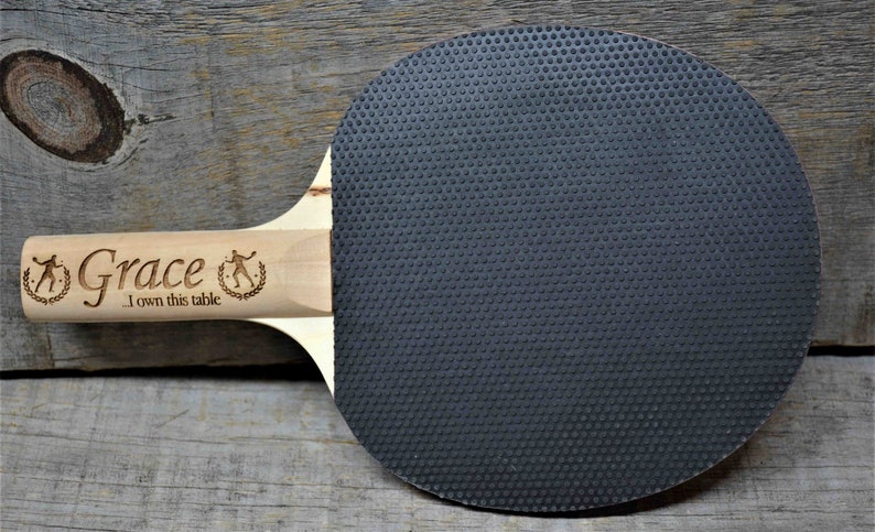 May include: A wooden ping pong paddle with a black rubber surface. The handle is engraved with the name "Grace" and the text "I own this table".