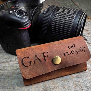 May include: A brown leather wallet with the initials "GAF" engraved on the front, along with the date "est. 11.03.67". The wallet has a brass snap closure. A black camera is in the background.