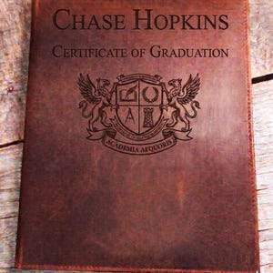 May include: A brown leather certificate holder with the name "Chase Hopkins" and "Certificate of Graduation" embossed on the cover. The holder features a detailed crest with two winged creatures and the text "Academia Aequoris".