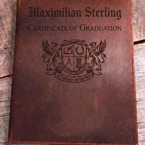 May include: A brown leather certificate holder. The cover is embossed with the name "Maximilian Sterling" and "CERTIFICATE OF GRADUATION". A crest featuring griffins and the words "ACADEMIA AEQUORIS" is also visible.