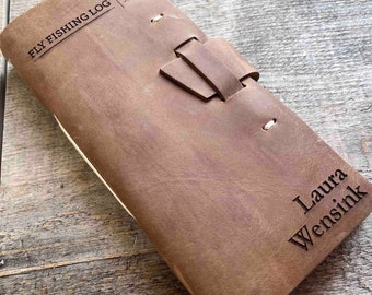 May include: A brown leather journal with a strap closure and the text "FLY FISHING LOG" embossed on the front cover. The journal is personalized with the name "Laura Wensink" on the back cover.