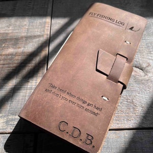 Puede incluir: Un libro de registro de pesca con mosca de cuero marrón con una cita: "Ten cuidado cuando las cosas se pongan difíciles y nunca te des la vuelta." El libro tiene un cierre de correa de cuero y las iniciales C.D.B. están grabadas en la parte delantera.