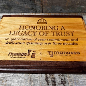 Puede incluir: Una placa de madera con un borde marrón, grabada con el texto "Honoring a Legacy of Trust" y los logotipos de Franklin Adhesives & Polymers y TJ ManosSO Representações. La placa es un reconocimiento al compromiso y la dedicación que se extiende por más de tres décadas.
