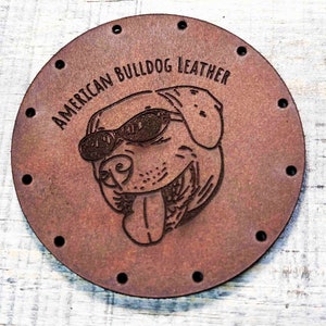 Peut inclure: Un patch en cuir marron avec un bouledogue de dessin animé portant des lunettes de soleil et tirant la langue. Le texte "AMERICAN BULLDOG LEATHER" est imprimé sur le patch.