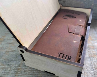 May include: A brown leather-bound journal with the words "Fishing Log" and a fly fishing hook and fly design engraved on the front cover. The journal is in a wooden box with the initials "THR" engraved on the front.