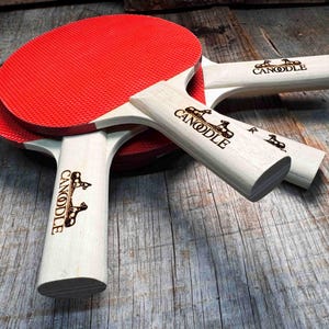 May include: Two red and black ping pong paddles with wooden handles. The handles are engraved with the word "CANOODLE" and a logo of two horses.