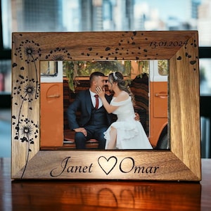 May include: A wooden photo frame with a brown finish. The frame has a dandelion design carved into the wood. The frame is holding a photo of a couple in front of a vintage van. The photo is dated 13.01.2024. The names Janet and Omar are engraved on the frame with a heart symbol between them.