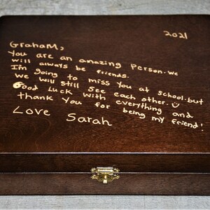 May include: A dark brown wooden box with a gold latch. The top of the box is inscribed with a handwritten message that reads: "2021 Graham, You are an amazing Person. We will always be friends. I'm going to miss you at school. But we will still see each other. Good luck with everything and thank you for being my friend! Love Sarah"